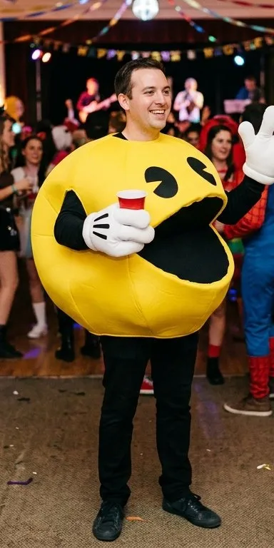 Hombre con disfraz de comecocos amarillo, un cuerpo redondo con la boca y los ojos del personaje impresos, mangas negras y guantes blancos, con un vaso de fiesta en la mano