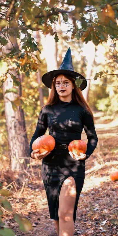 Mujer disfrazada de bruja con vestido negro, sombrero de pico y dos calabazas en las manos, en un bosque