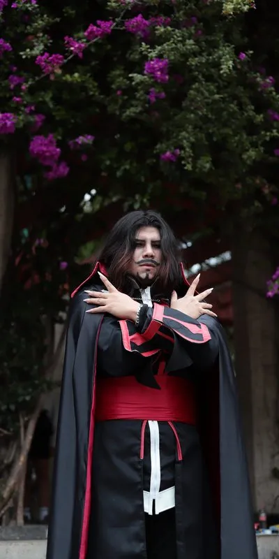 Hombre disfrazado de vampiro con capa negra de forro rojo, túnica oscura con fajín rojo y uñas largas blancas