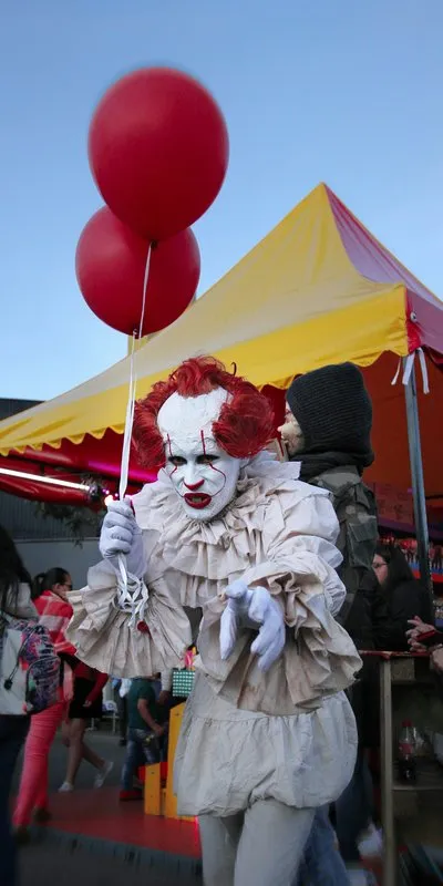 Persona disfrazada de payaso siniestro con maquillaje blanco, pelo rojo rizado, cuello de gorguera y dos globos rojos en la mano