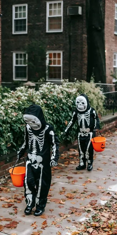 Dos niños disfrazados de esqueleto con mono negro, capucha y máscara, sosteniendo cubos de calabaza para caramelos en una calle con hojas de otoño