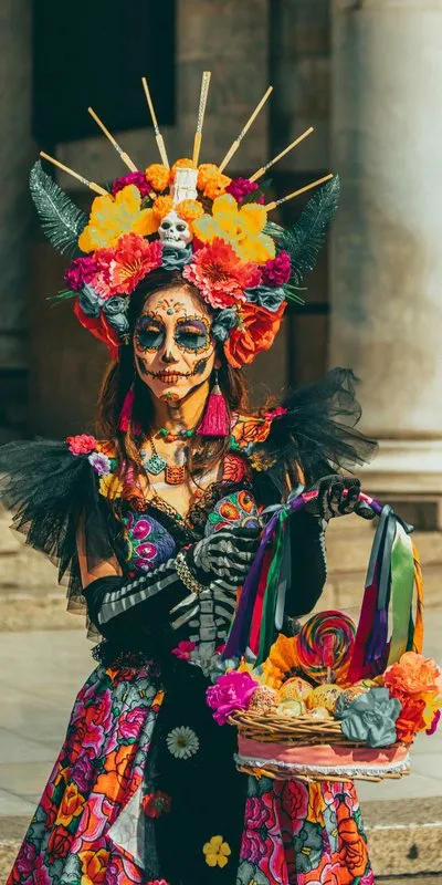Mujer disfrazada de Catrina con maquillaje de calavera floral, tocado de flores y vestido de colores, sosteniendo una cesta