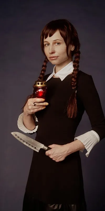 Mujer disfrazada de Wednesday Addams con vestido negro de cuello blanco, dos trenzas, sosteniendo una vela roja y un cuchillo