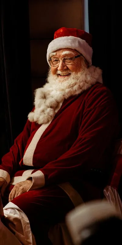 Hombre mayor disfrazado de Papá Noel, sentado en un sillón, con chaqueta roja de terciopelo y ribete blanco de pelo, gorro rojo con pompón, gafas y barba blanca larga, sonriendo