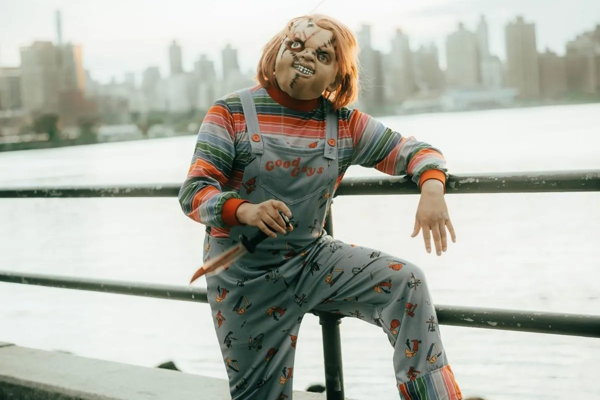 Persona disfrazada de Chucky con máscara de muñeco cicatrizado, peluca pelirroja, camiseta de rayas de colores y peto vaquero con el logotipo Good Guys