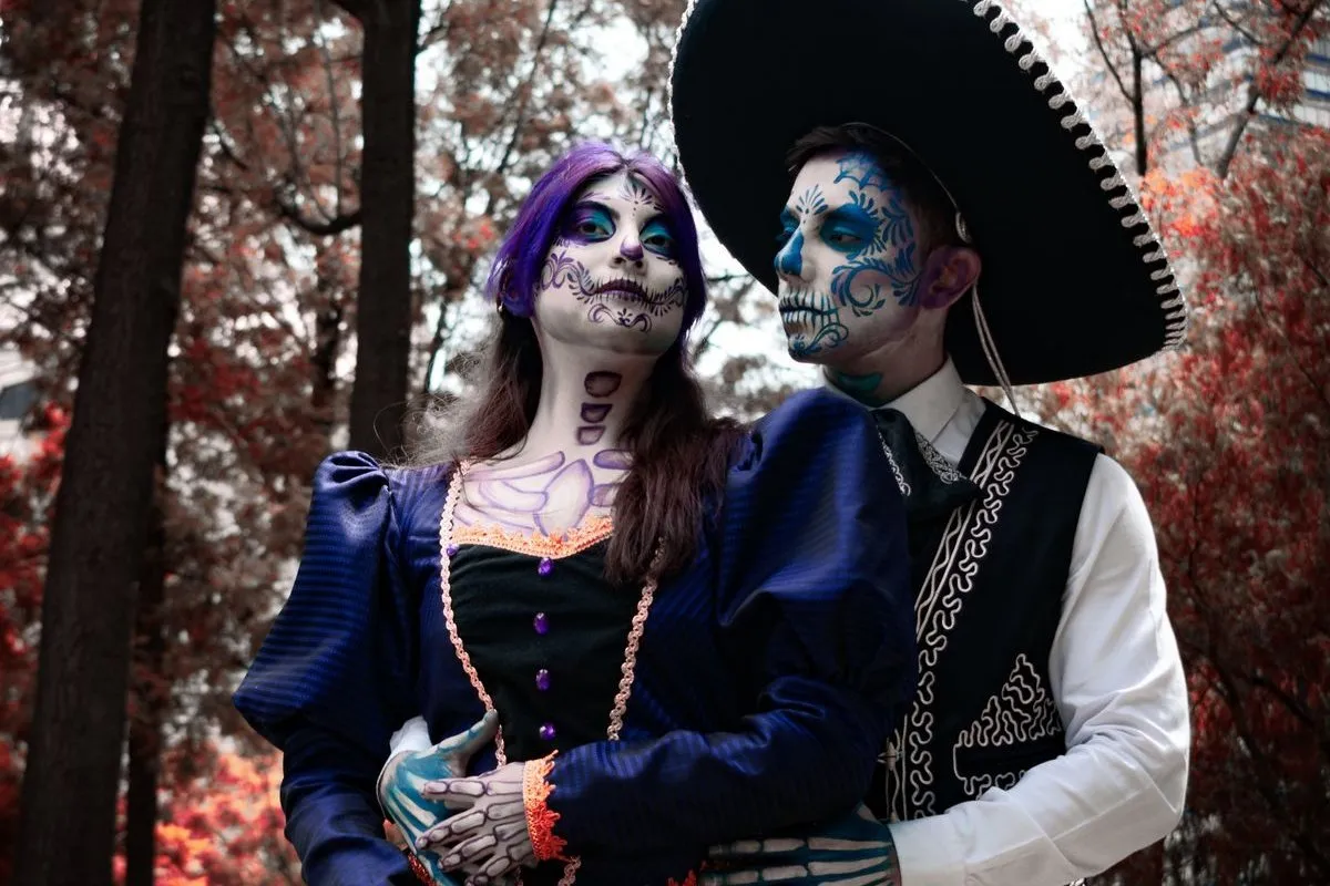 Pareja con maquillaje de calavera mexicana, ella con vestido azul de encaje y guantes pintados de huesos, él con sombrero de charro y chaleco bordado